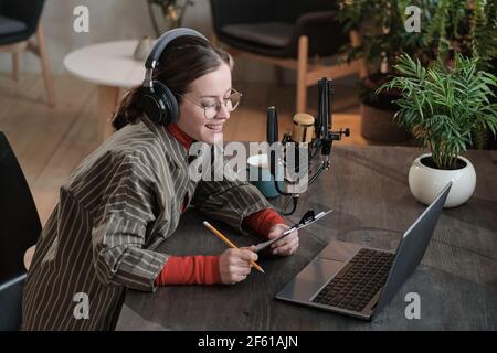 Female journalist sitting at the table in front of laptop and doing online interview on it in the studio Stock Photo