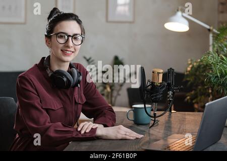 Portrait of young podcaster in headphones smiling at camera while sitting at the table in front of laptop and working in radio studio Stock Photo