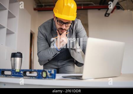 Focused architect looking at blueprints and thinking while leaning on desk at construction site. Stock Photo