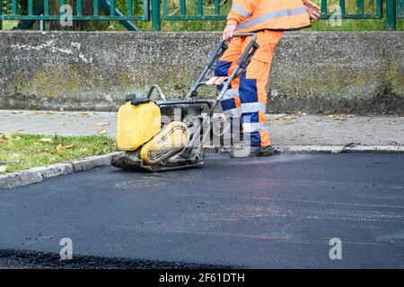 Paving worker uses vibratory plate compactor to compact new asphalt near curbstones Stock Photo
