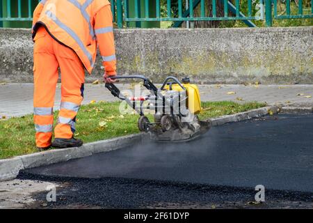 Paving worker uses vibratory plate compactor to compact new asphalt near curbstones Stock Photo
