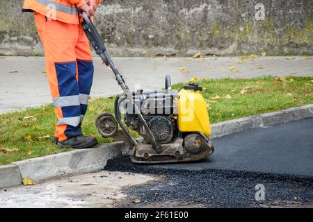 Paving worker uses vibratory plate compactor to compact new asphalt near curbstones Stock Photo