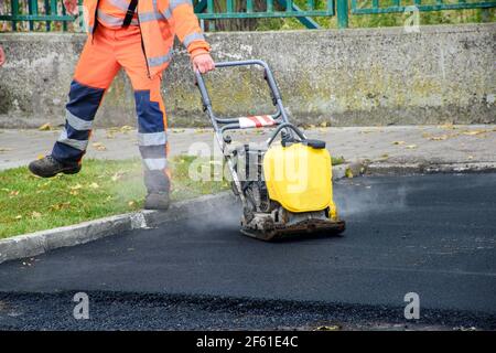 Paving worker uses vibratory plate compactor to compact new asphalt near curbstones Stock Photo