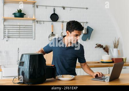 Handsome young caucasian man looking at instructions online on laptop on how to use modern air fryer to cook healthy food without using oil at home in kitchen Stock Photo