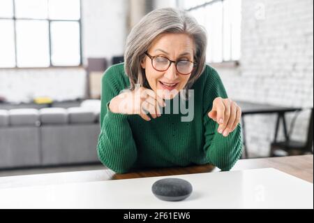 Contemporary retired woman using voice commands to control smart speaker, mature woman talking to the digital virtual assistant at home, asking a question or requesting to switch music Stock Photo
