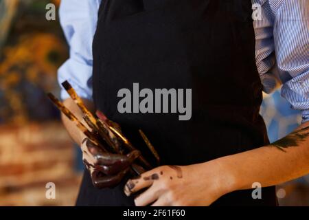 Close up view of female artist hands stained with paint in art studio. Painter wearing apron holding art brushes. Creative process, relaxation Stock Photo