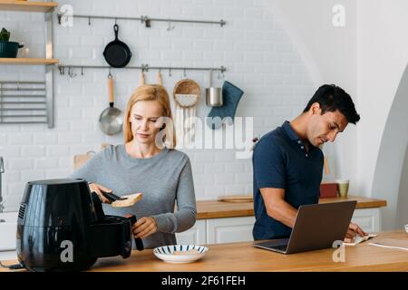 Beautiful young caucasian wife in kitchen cooking healthy oil free meal in modern air fryer with handsome husband working on laptop in kitchen at home Stock Photo