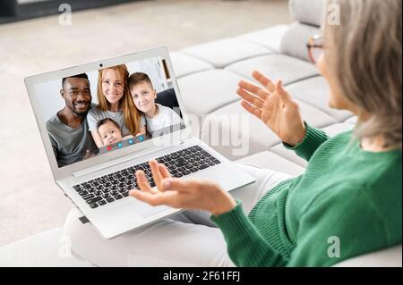 Back view a grey haired senior woman in touch with a multiracial family of her grown kids, grandmother using laptop computer for video call to grandchildren, video meeting Stock Photo