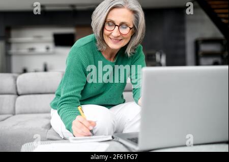 Confident mature woman using laptop computer for remote work, watching webinar and taking notes sitting at home. Contemporary senior female online teacher holding video conference and making marks Stock Photo
