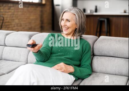 Cheerful senior woman enjoys evening with a TV shows, series. Smiling mature woman sits on the sofa with TV remote controller and switching channels Stock Photo