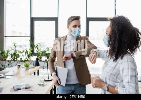 interracial business partners in medical masks doing elbow greeting in office Stock Photo