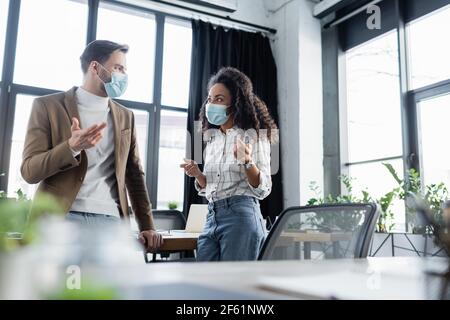 young interracial business people in protective masks gesturing while talking in office, blurred foreground Stock Photo