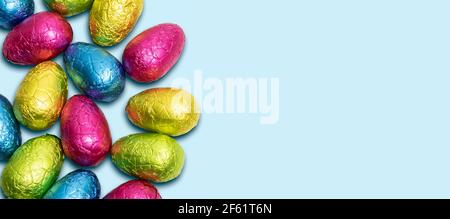 Side border of colorful wrapped chocolate Easter eggs on blue background. Top view. Stock Photo