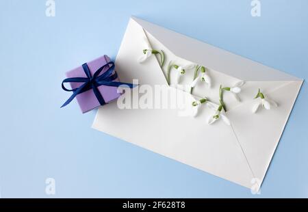 Beautiful snowdrops in white envelope and purple gift box on light blue background, closeup, space for text, top view. Stock Photo