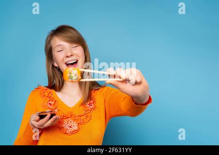 Enlarged photo of a happy blonde girl who with a smile holds sushi with wooden chopsticks and a bowl with sauce. Blue background and side space. Stock Photo