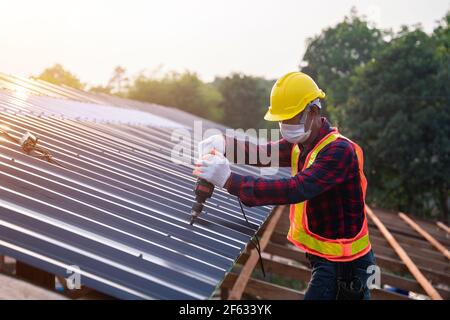 Asian construction worker safety wear using electric drill tools install on new roof metal sheet, Roof construction concept. Stock Photo