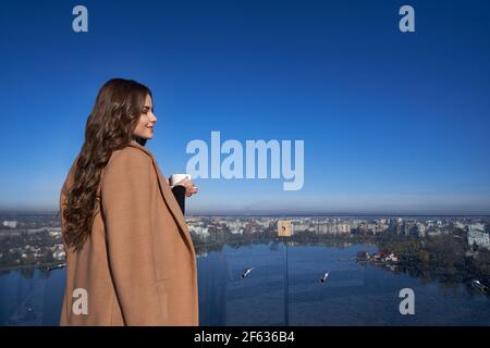 Attractive young woman drinking morning coffee while standing balcony. Pleasant lady enjoying beautiful view on big city. Concept of relaxation. Stock Photo