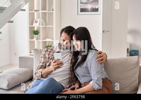 Smiling indian family couple using smartphone ordering online on couch at home. Stock Photo
