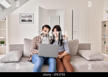 Happy indian couple using laptop computer sitting on sofa together at home. Stock Photo