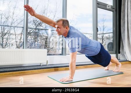 Middle age man stay on floor mat in plank pose and stretching hands at light studio background, healthy fitness yoga exercises. Plank with arm lift. Stock Photo