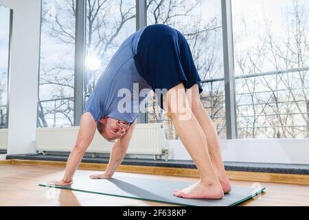 MIddle age man stay on floor mat, making workout yoga training in downward dog pose, tild and bend body muscles at light fitness studio Stock Photo