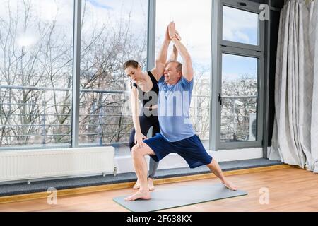 Middle age man make stretching workout exercises on mat in light fitness studio with panoramic window and female instructor assistance. Warrior pose w Stock Photo