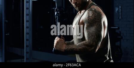 Portrait of muscular tattooed incognito bodybuilder doing crossover exercise. Side crop of bearded man with perfect body training triceps in gym in dark atmosphere. Concept of bodybuilding. Stock Photo