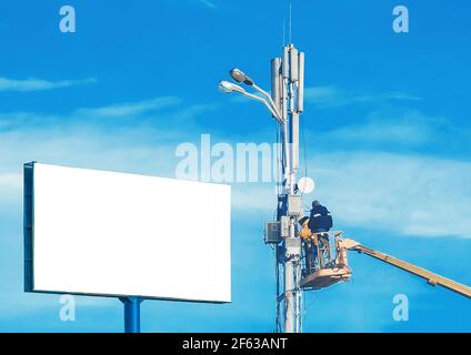 A mock-up large white billboard form and two construction workers on a machine crane repair a lighting pole against a blue sky. Stock Photo