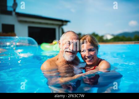 Cheerful senior couple in swimming pool outdoors in backyard, looking at camera. Stock Photo