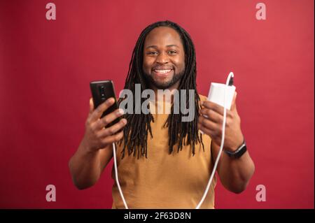 Happy satisfied African American guy with dreadlocks standing isolated on red wall, holding mobile phone connected to power bank through USB cable, charging his device, smiling and looking at camera Stock Photo