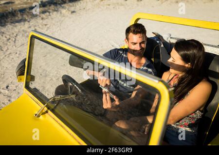 Happy caucasian couple sitting in beach buggy by the sea using smartphone Stock Photo