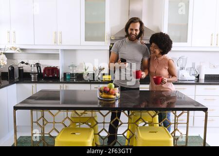 Happy diverse couple standing in kitchen drinking coffee and using smartphone Stock Photo