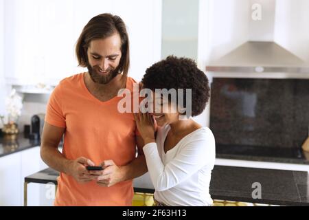 Happy diverse couple in kitchen using smartphone and embracing Stock Photo