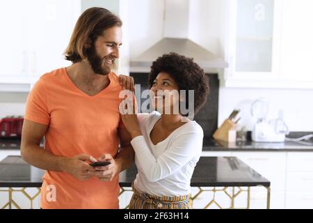 Happy diverse couple in kitchen using smartphone and embracing Stock Photo