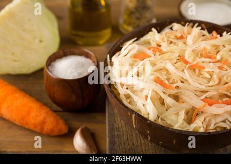 Close up view of part of bowl of sour sauerkraut on brown, wooden background. Next to is sliced fresh cabbage and carrots, salt, garlic and olive oil. Stock Photo