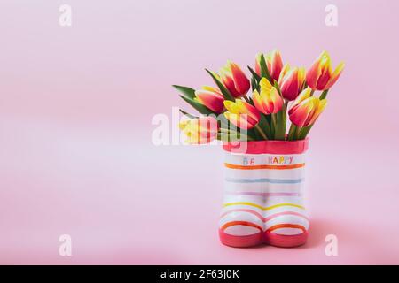 Fresh red and yellow tulips in a vase with the shape of children colorful boots with Be happy lettering on pastel pink background, Inspiration card Stock Photo