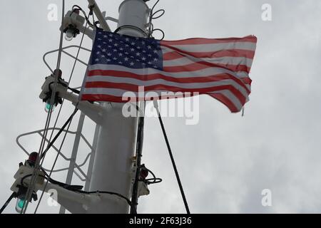 American flag on the wind hanging on navigational mast with green lights of the bridge of merchant container vessel during mooring in American port. Stock Photo