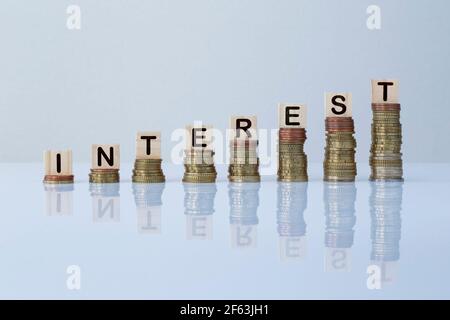 Word 'INTEREST' on wooden blocks on top of ascending stacks of coins against gray background. Concept photo of economy, business, finance and banking. Stock Photo