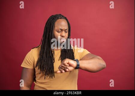 Serious confused young African American man perfectionist with dreadlocks isolated on red looking and checking time from watch on his wrist, waiting too long, time is up or deadline concept Stock Photo
