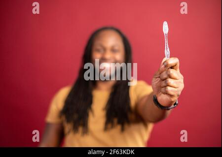 Selective focus at the toothbrush in hand of smiling man. Happy African American man with dreadlocks and great smile on red background, advertising toothbrush, toothpaste, hygiene concept Stock Photo