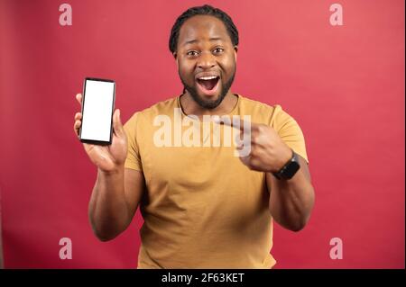 Surprised African-American man holds smartphone in a hand and points finger at empty phone screen, astonishment black guy presenting best deal, advertising new mobile app, isolated on red Stock Photo