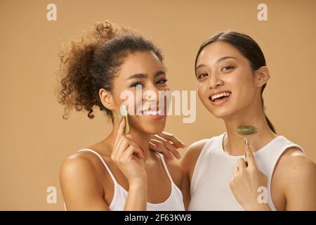 Pretty young women smiling at camera, using jade roller and facial gua sha while posing together isolated over beige background Stock Photo