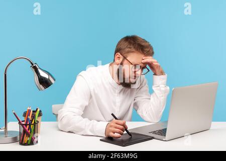 Serious busy man editor with beard in eyeglasses working on graphic tablet, drawing sketches or retouching photos, sitting at workplace with laptop. I Stock Photo