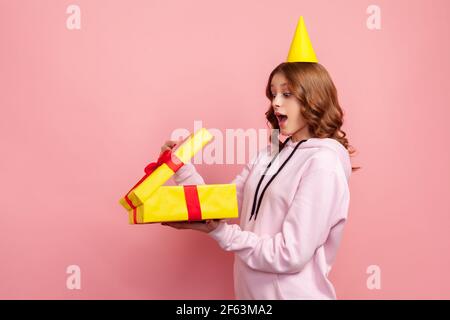 Profile of amazed happy teenage girl in hoodie opening gift box with opened mouth, satisfied with present. Indoor studio shot isolated on pink backgro Stock Photo