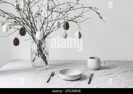 Easter festive table setting. Fresh branches in vase decorated by paper ornaments. Hanging chicken eggs with quail festher.Spring interior decoration Stock Photo