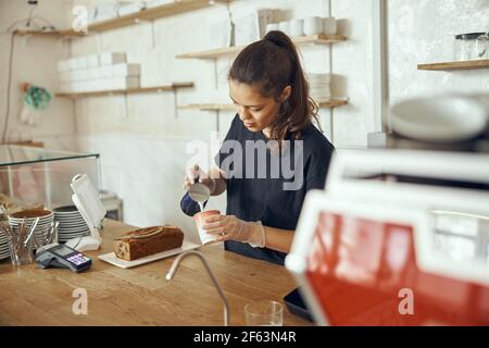 Young female woman barista pouring fresh milk to prepare latte coffee for customer in cafe. For small business startup in food industry concept Stock Photo