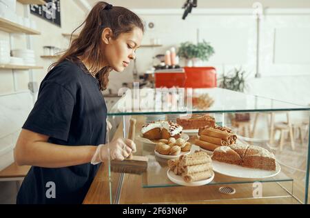 Female barista at showcase with desserts in cafe. Young worker of sweet shop and cafe Stock Photo