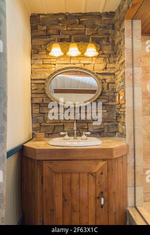 Pine wood vanity with white porcelain sink and antique faucets, oval mirror and three bell-shaped lighting fixtures in opened design bathroom Stock Photo