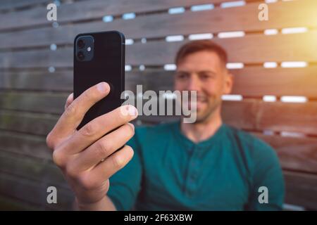 Young happy man with smartphone outdoors at sunset. Man using modern black smartphone. Man having a facetime video call. Looking at smartphone  Stock Photo