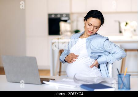 A tired overworked mixed race pregnant adult woman, designer, manager or freelancer working remotely at desk, taking a break, have a back pain, does back massage with hands Stock Photo